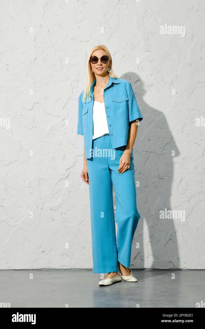 Femme souriante dans des lunettes de soleil et tenue de printemps à la mode bleue debout près d'un mur Banque D'Images