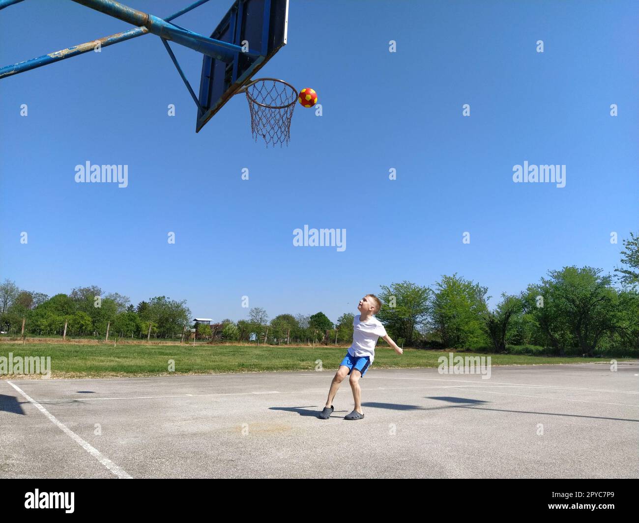 Sremska Mitrovica, Serbie. 6 juin 2020. Un garçon joue au ballon sur le terrain de jeu. Terrain de sport en asphalte. Un enfant dans un t-shirt blanc. Enfant en bas âge aux cheveux blonds, 7 ans. Courir, donner des coups de pied et faire des exercices Banque D'Images