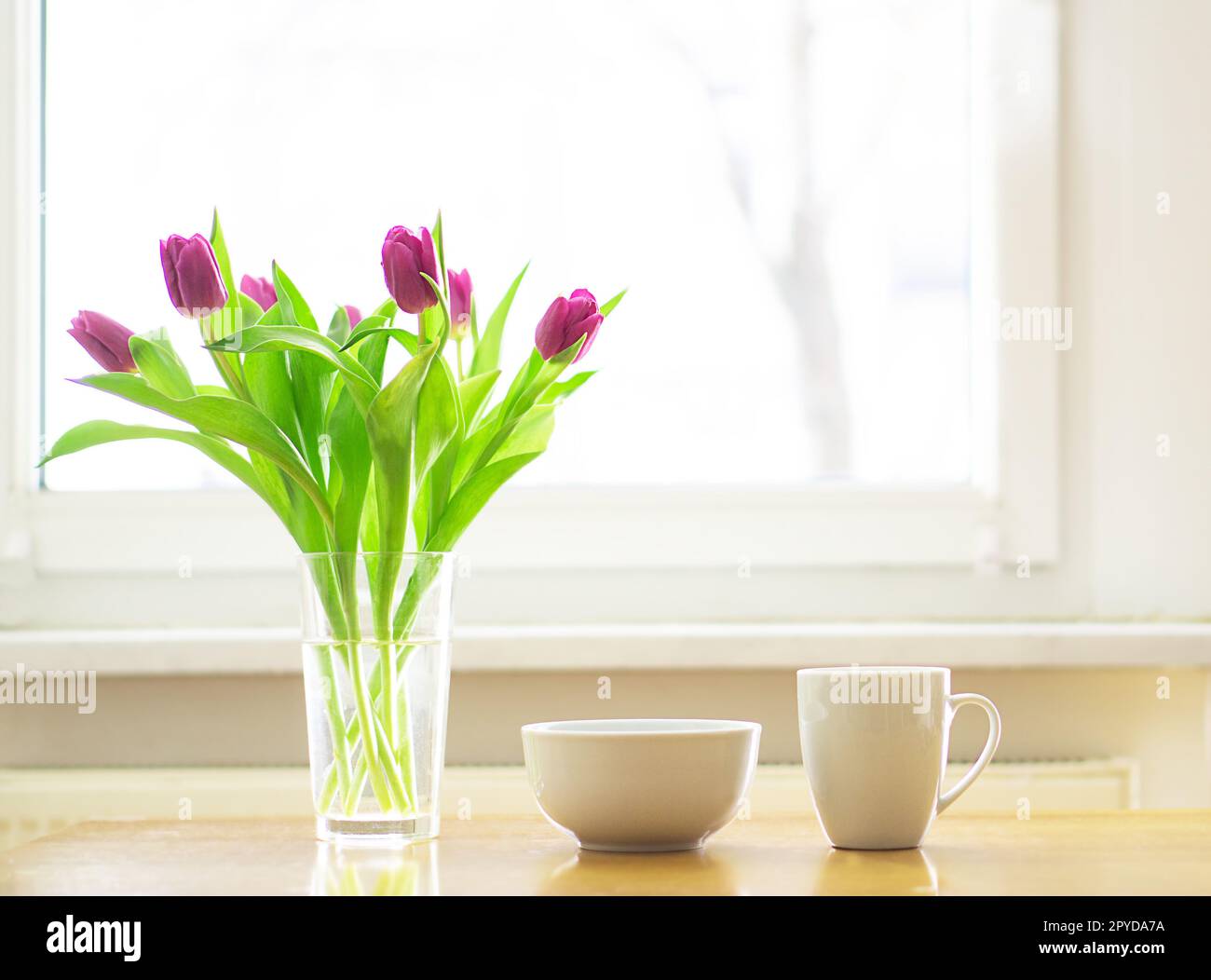 Petit déjeuner confortable avec une tasse de thé chaud ou de café et un bol de muesli avec un bouquet de tulipes magenta sur la table en bois devant une fenêtre. Bonjour. Espace de copie. Photo de haute qualité. Banque D'Images