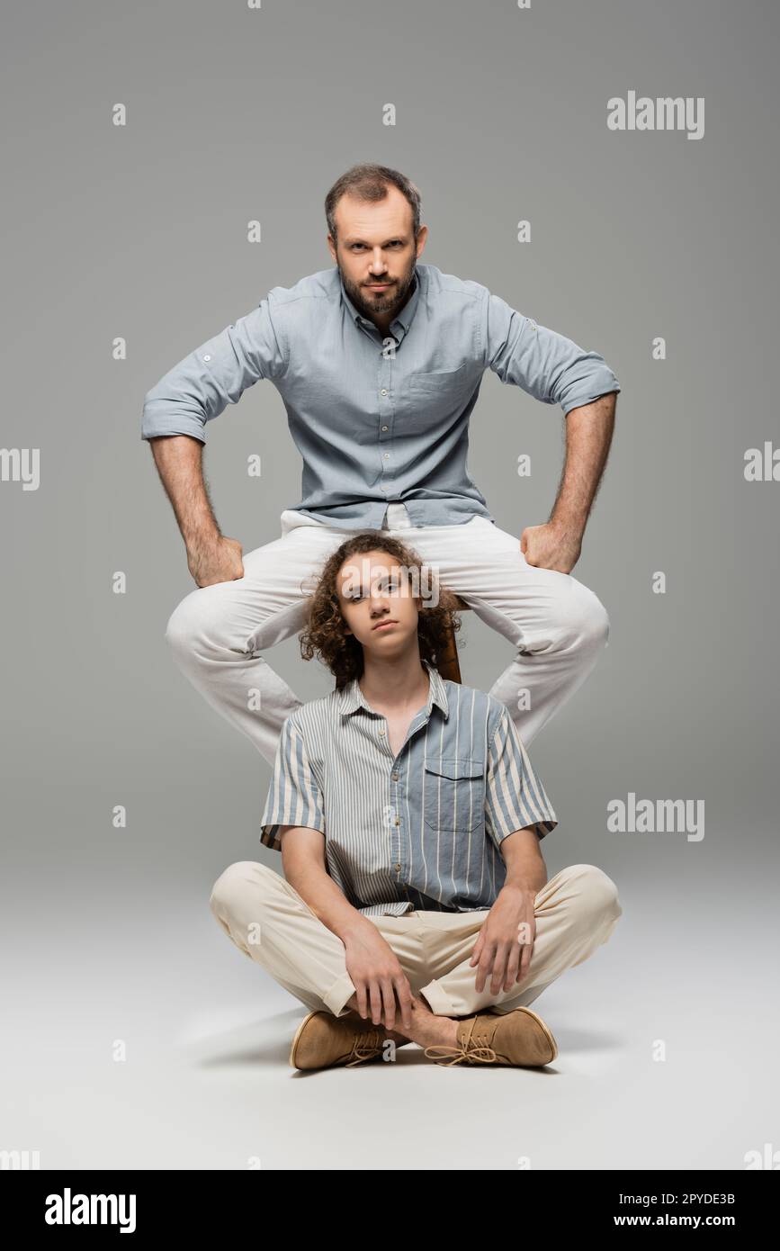 papa puissant et barbu assis sur une chaise à côté de l'adolescent bouclé garçon sur gris, image de stock Banque D'Images