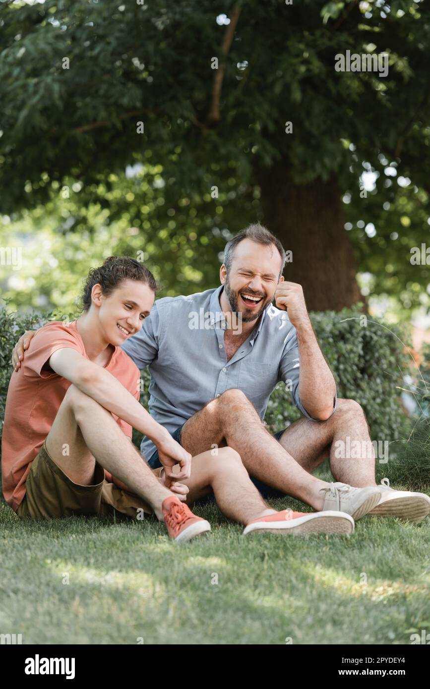 père et fils joyeux assis dans des vêtements d'été sur une pelouse verte dans le parc, image de stock Banque D'Images