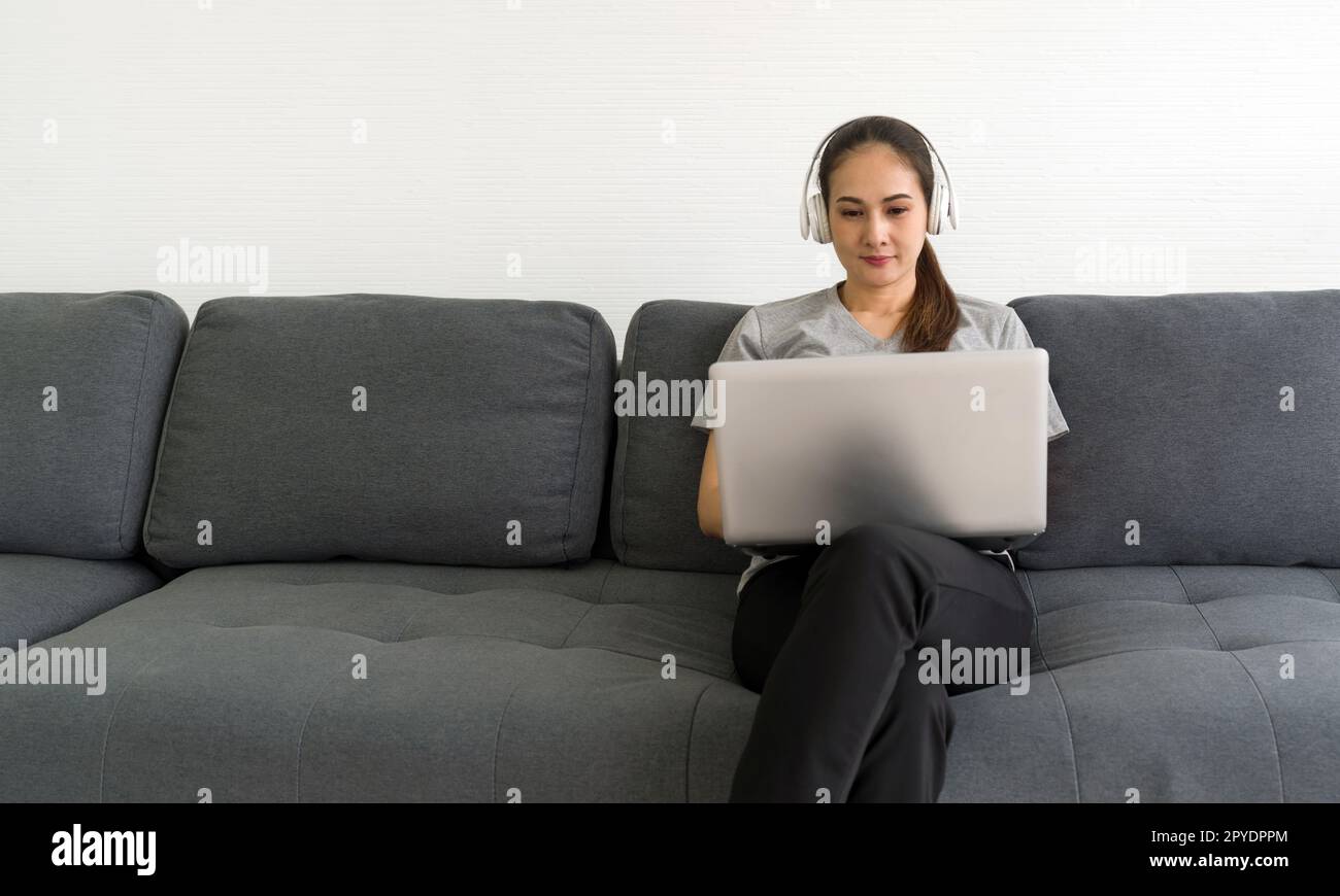 Femme asiatique d'âge moyen en t-shirt gris écouter de la musique à partir d'un casque tout en tapant sur un ordinateur portable dans le salon, profiter d'un week-end de loisirs à la maison. Concept sans stress Banque D'Images