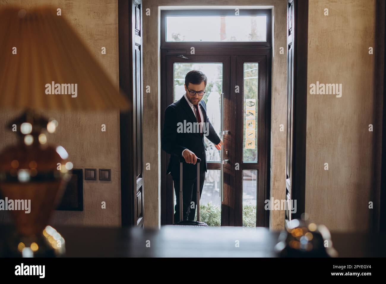 Agréable homme souriant en costume classique devant l'entrée d'un hôtel d'affaires traînant ses bagages Banque D'Images