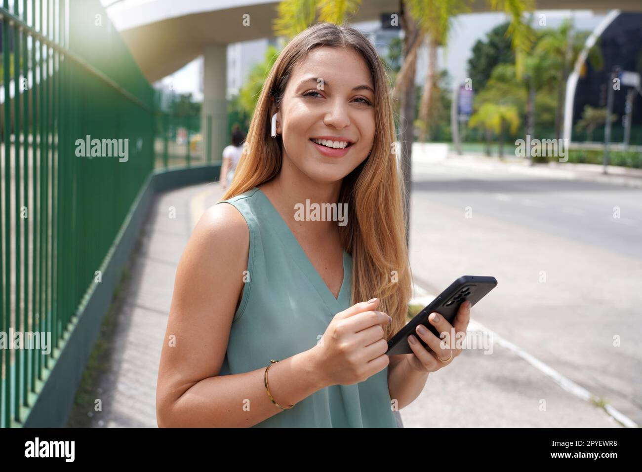 Femme brésilienne sourire à la caméra à l'aide d'un smartphone pour les médias sociaux dans la ville avec des écouteurs à l'extérieur. Heureuse étudiante latine souriante pour la technologie de connexion Internet 5G sur téléphone portable. Banque D'Images