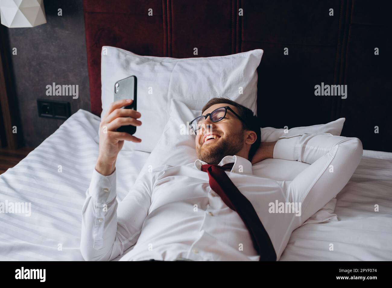 Un homme d'affaires se repose sur un lit dans une chambre d'hôtel. Un jeune homme utilise son smartphone pendant qu'il se repose. Banque D'Images