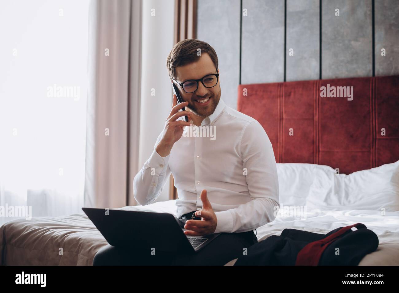 Beau homme d'affaires qui fait le téléphone tout en étant assis sur le lit de la chambre d'hôtel et en utilisant un ordinateur portable pendant un voyage d'affaires Banque D'Images