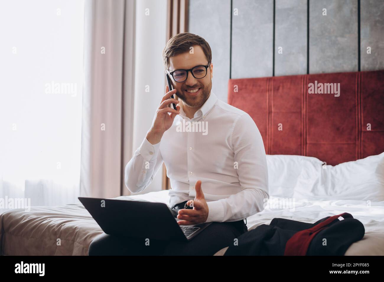 Beau homme d'affaires qui fait le téléphone tout en étant assis sur le lit de la chambre d'hôtel et en utilisant un ordinateur portable pendant un voyage d'affaires Banque D'Images