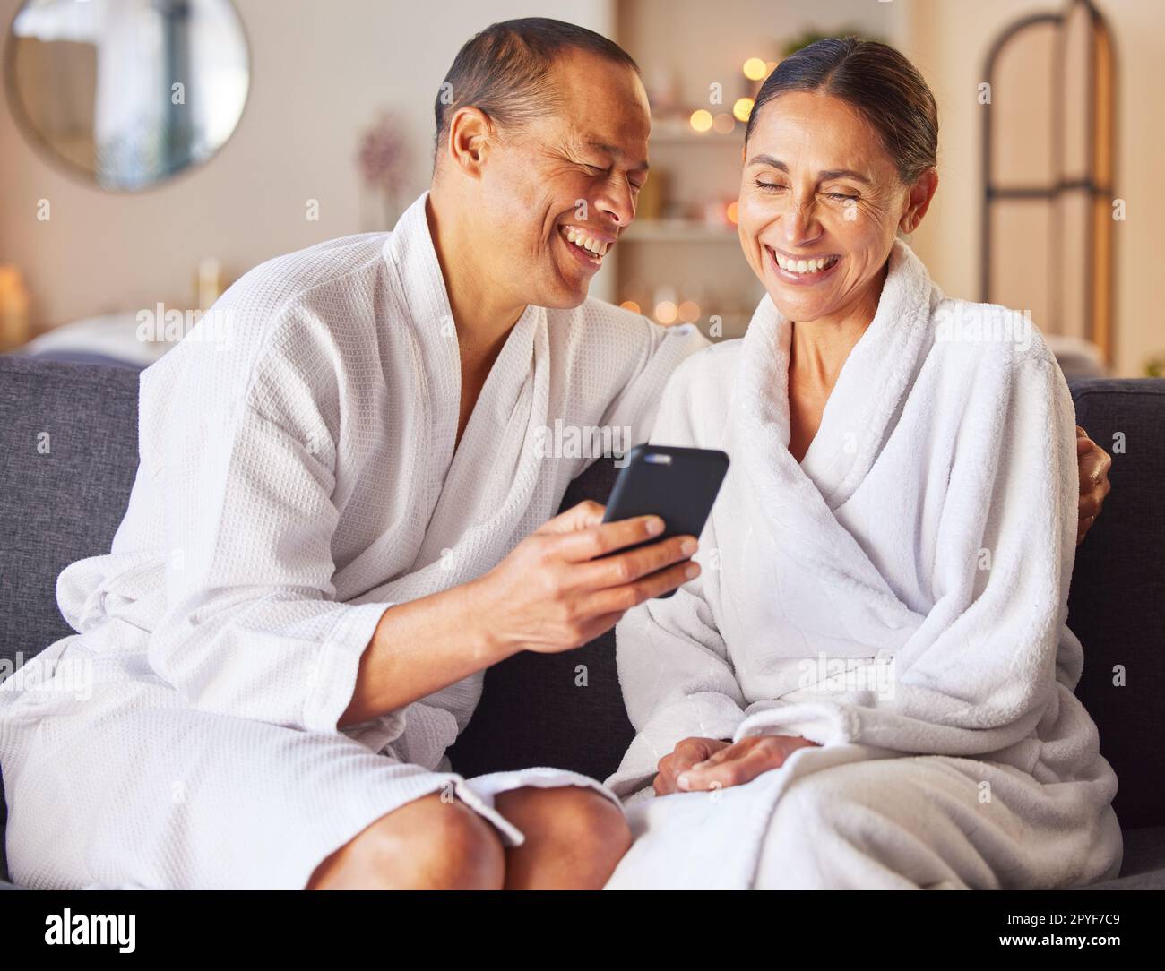 Couple senior, spa et se détendre heureux avec téléphone sur le canapé, centre de bien-être et soins du corps de luxe ou vidéo en streaming en ligne. Bonheur, femme et homme sourient ensemble, smartphone et robe de thérapie de beauté Banque D'Images