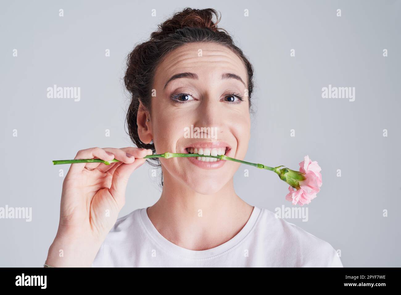 Ils n'ont jamais dit que ce n'était pas comestible. Photo studio d'une belle jeune femme tenant une fleur rose avec ses dents sur fond gris. Banque D'Images