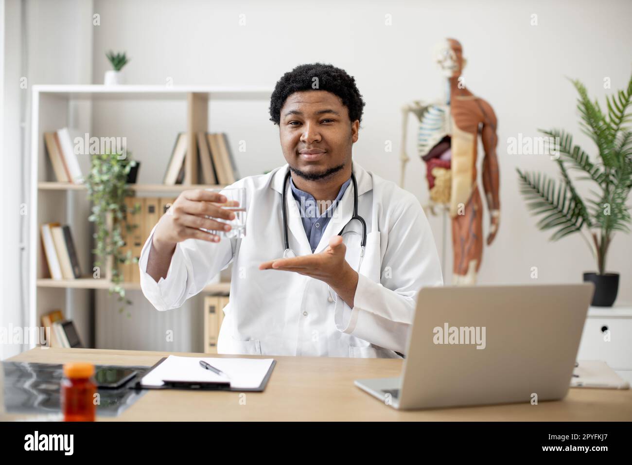 Médecin multiculturel positif en blouse de laboratoire montrant un verre d'eau tout en étant assis au bureau avec un ordinateur portable et des documents dans le bureau du médecin. Médecin généraliste recommandant de rester hydraté. Banque D'Images