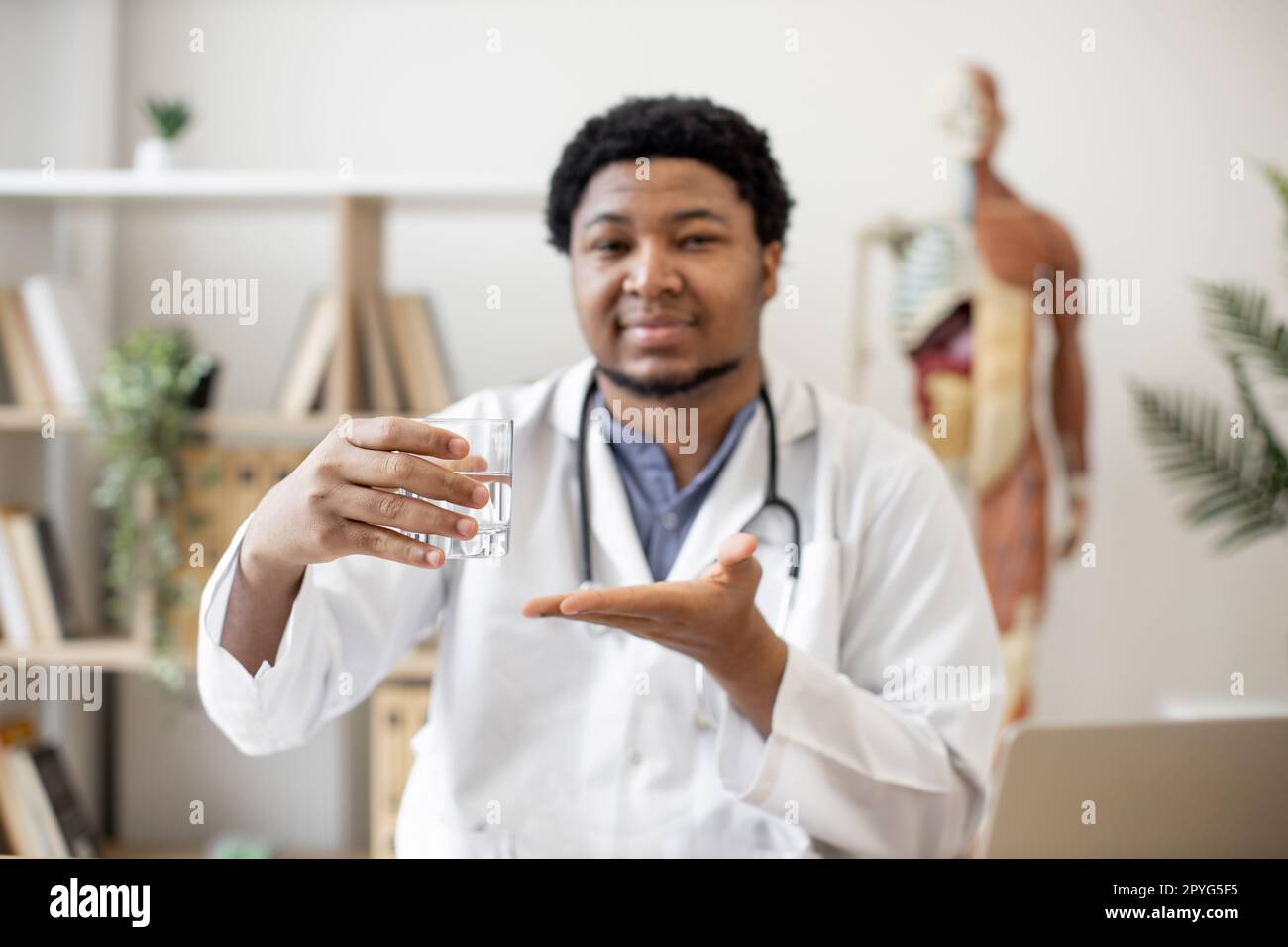 Médecin multiculturel positif en blouse de laboratoire montrant un verre d'eau tout en étant assis au bureau avec un ordinateur portable et des documents dans le bureau du médecin. Médecin généraliste recommandant de rester hydraté. Banque D'Images