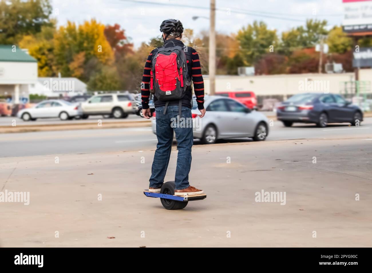Homme avec casque et dos sur une roue de skateboard motorisée sur le trottoir dans la zone urbaine avec des voitures sur la rade et les magasins et les arbres d'automne en arrière-plan - Banque D'Images