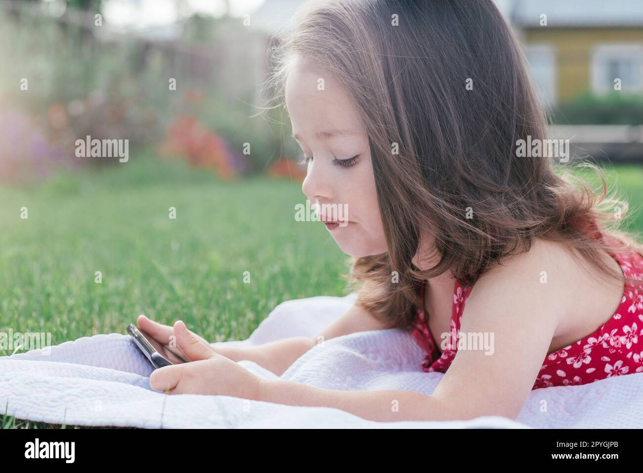 Portrait de petite fille 3-4 en rouge couché sur la couverture sur l'herbe verte et regardant dans le téléphone portable. Enfants utilisant des gadgets Banque D'Images