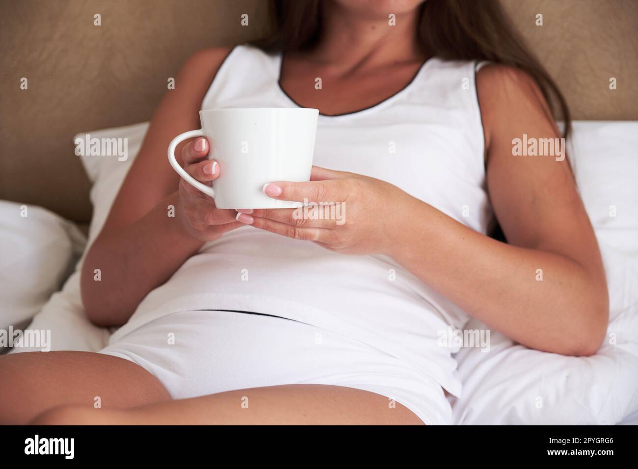 Tasse blanche dans les mains d'une jeune femme assise sur le lit dans la chambre le matin. Banque D'Images