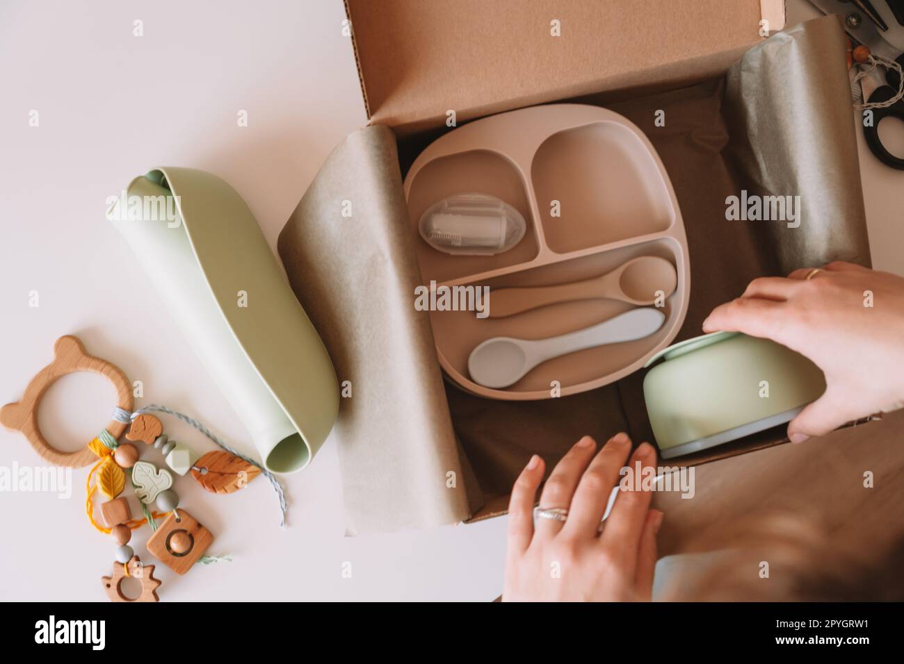 Mains recadrées emballage, emballage de l'expédition avec des plats de bébé en silicone, assiette, cuillères, bol, anneau de dentition écologique dans la boîte. Vue de dessus Banque D'Images