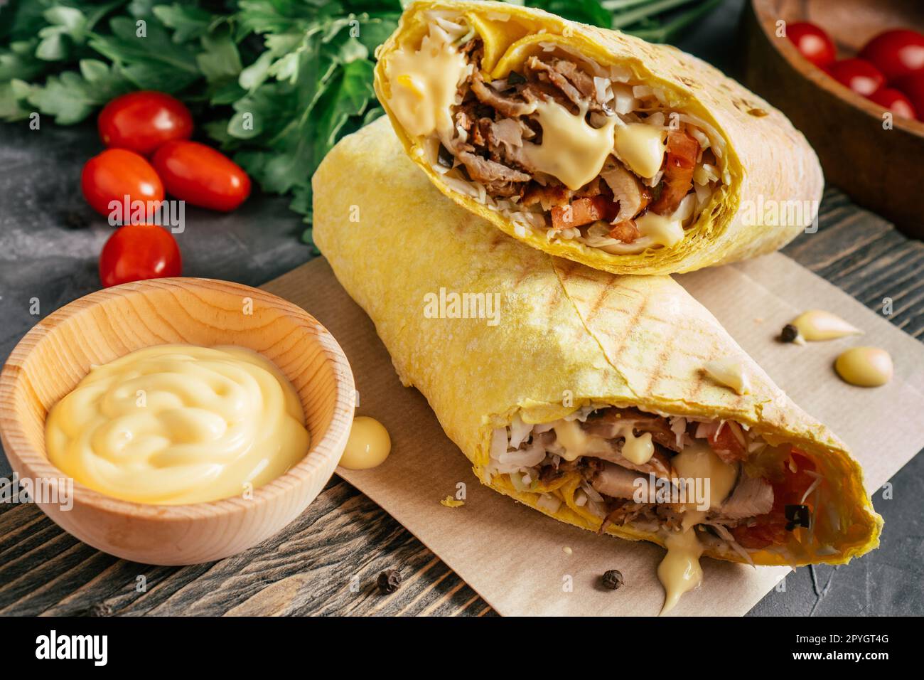 Shawarma au fromage avec sauce au fromage, poulet et légumes en pain pita sur surface en bois, tomates cerises et persil Banque D'Images