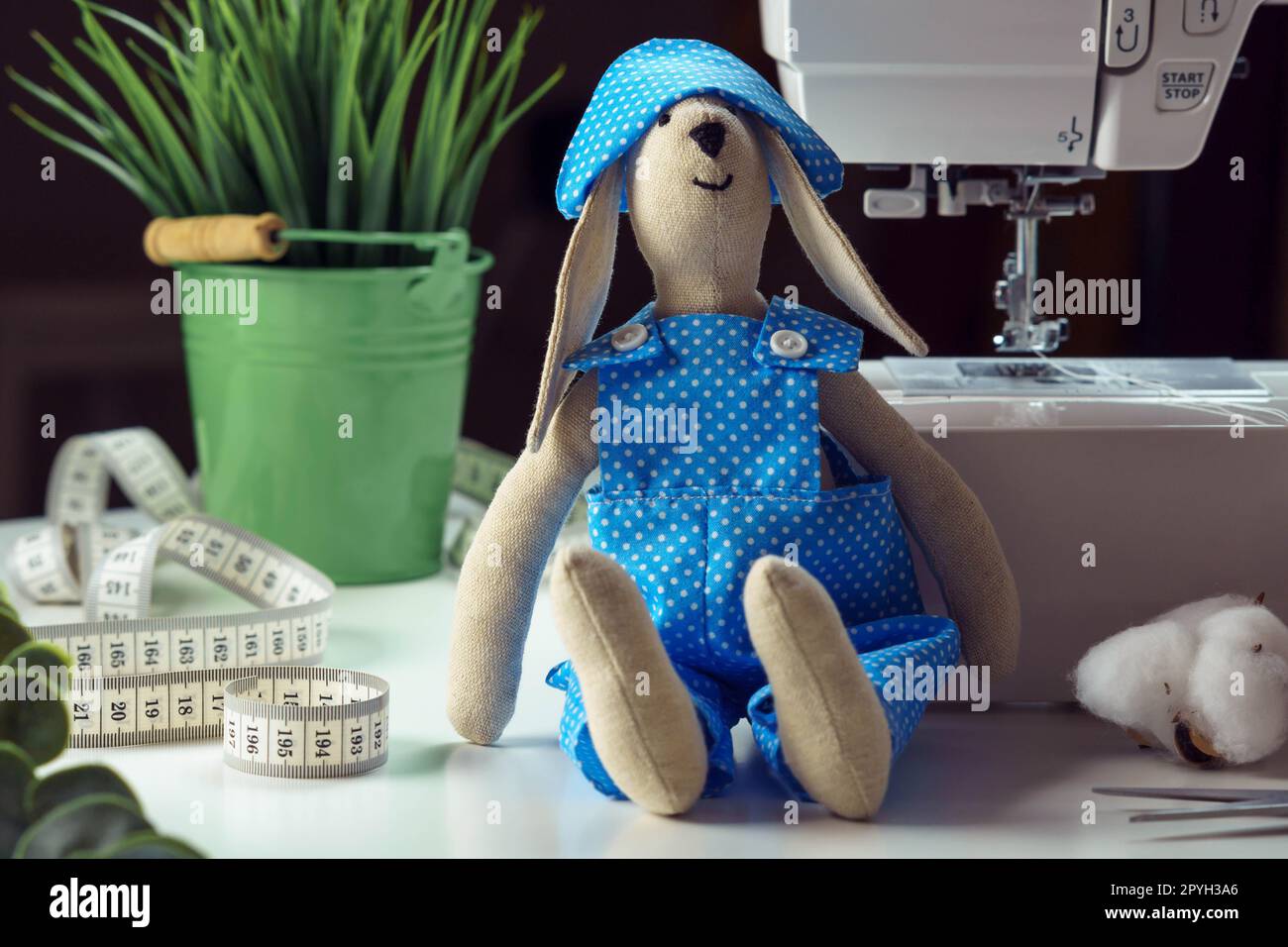 Mignon jouet lapin cousu en costume de design bleu assis près de la plante d'intérieur, ruban à mesurer et coton, penché à la machine à coudre Banque D'Images