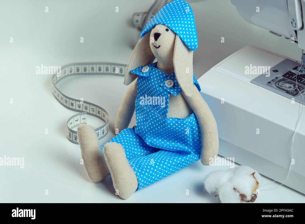 Jouet de lapin rembourré cousu souriant dans le costume de design bleu et chapeau assis près du ruban à mesurer, coton, maigre à la machine à coudre Banque D'Images