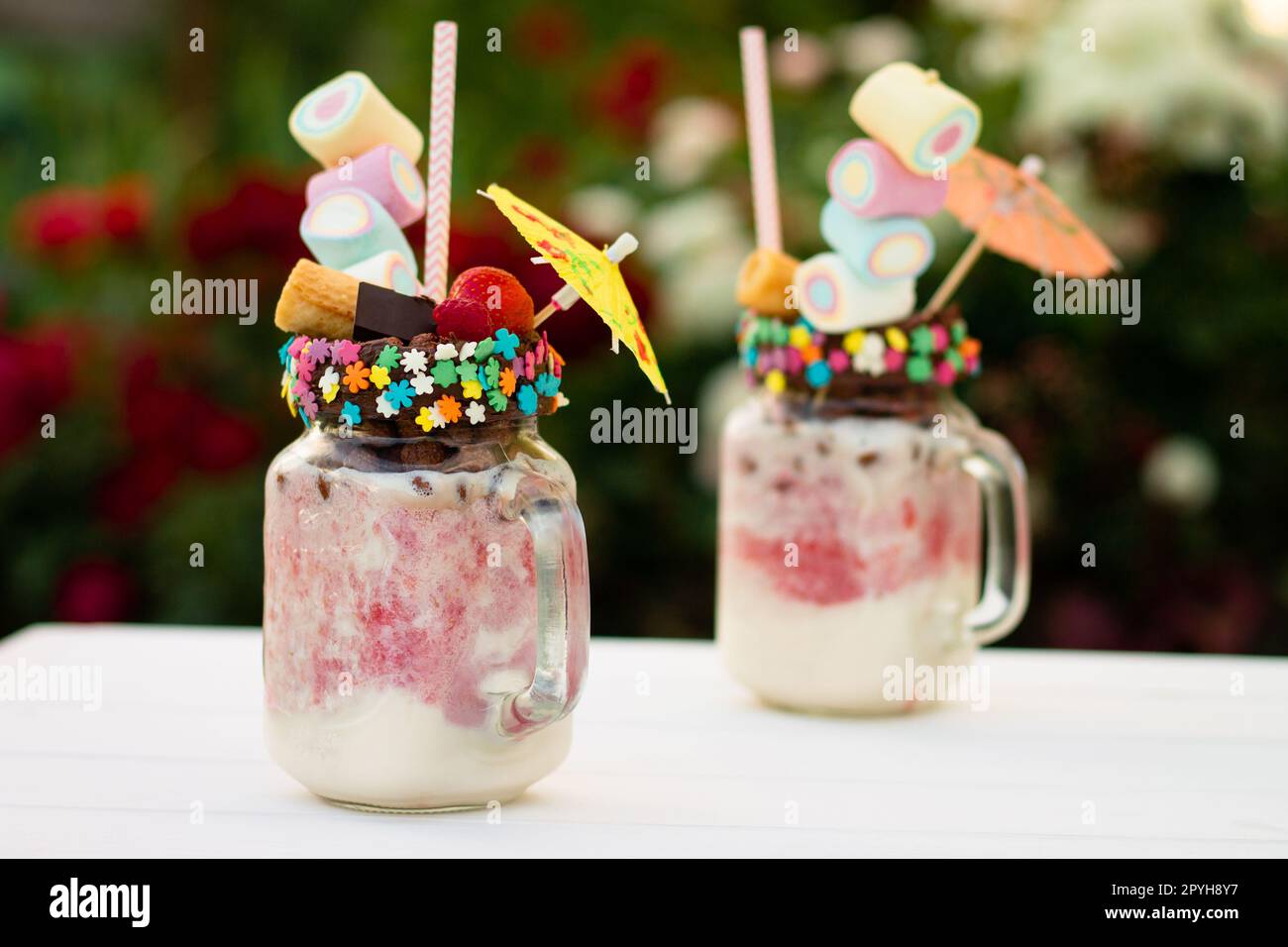 Freakshakes en pots avec paille, guimauve, fraise, chocolat sur une table blanche sur fond de fleurs. Banque D'Images