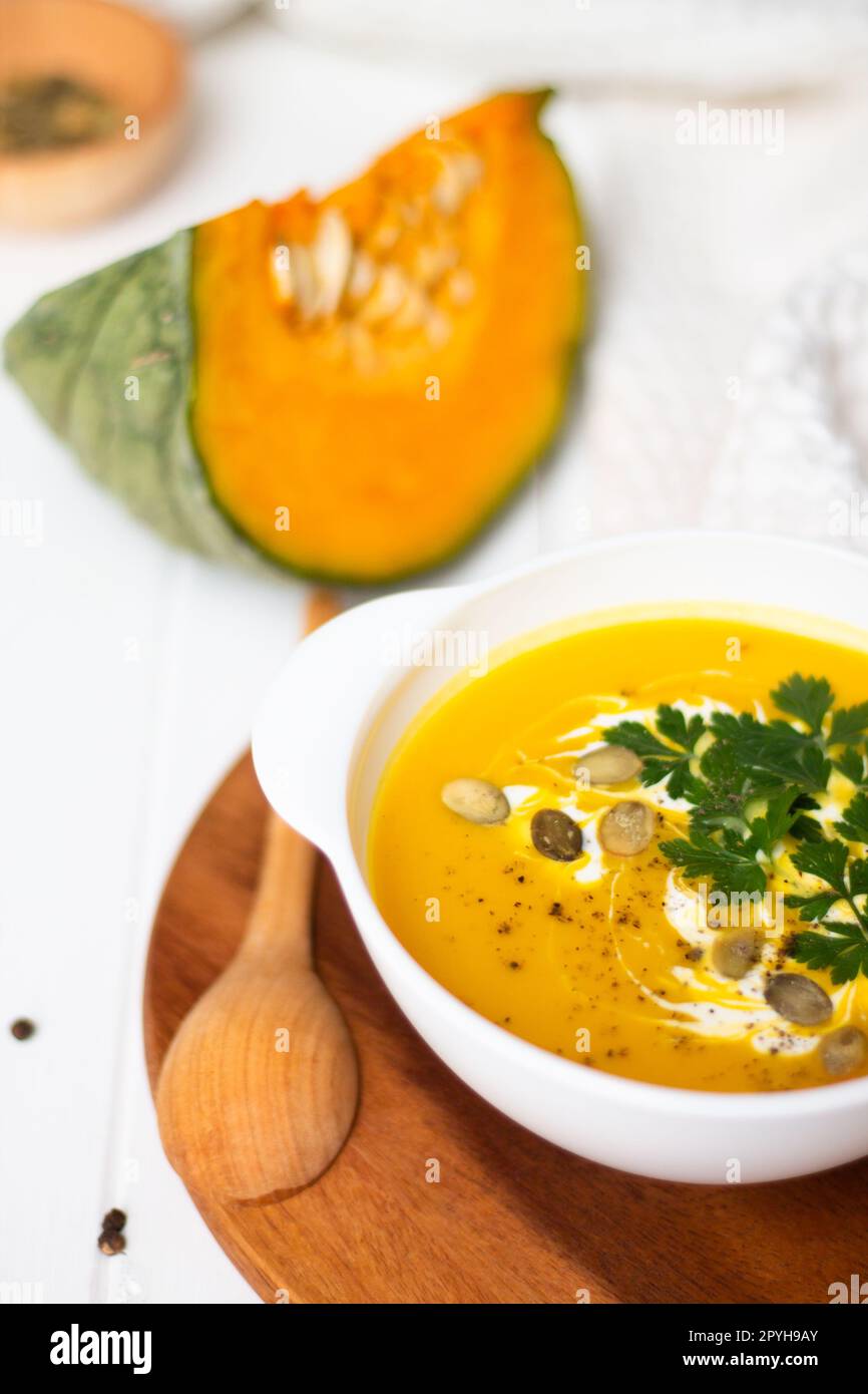 Une assiette de soupe à la crème de citrouille jaune avec de la crème, des herbes et des graines sur une table blanche près de la citrouille brute et d'une cuillère en bois. Banque D'Images