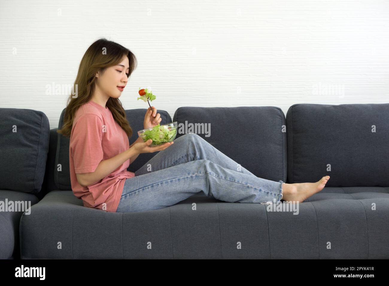Une jeune femme asiatique en t-shirt rose et jean s'assoient sur un canapé gris dans la salle de séjour, mangeant de la tomate et de la laitue tout en tenant un saladier. Banque D'Images
