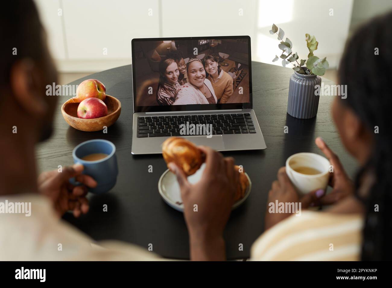 Vue arrière d'un jeune couple assis à une table devant l'ordinateur portable et prenant le petit déjeuner tout en ayant des appels vidéo avec des amis Banque D'Images