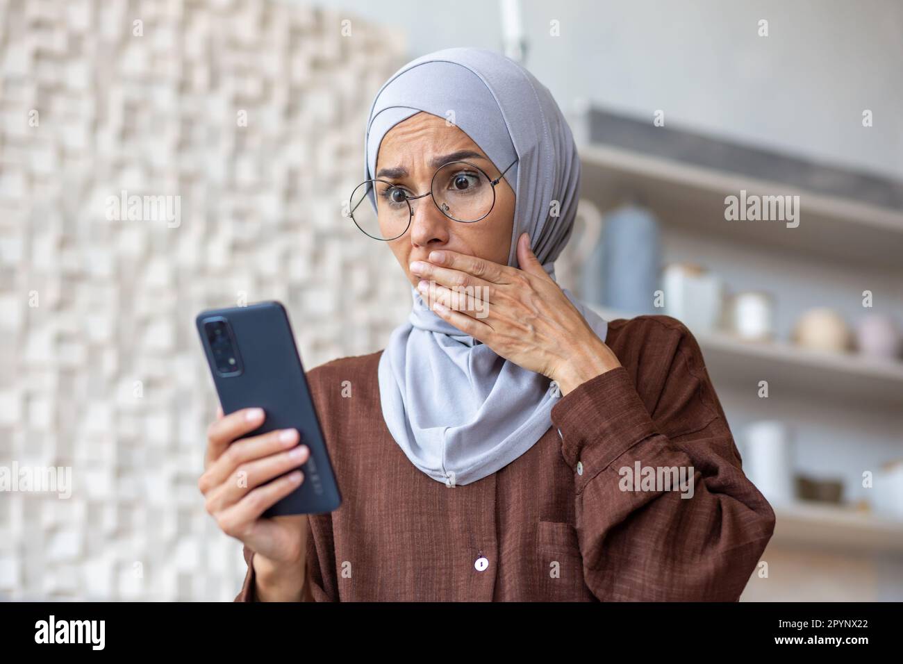 Jeune femme arabe dans le hijab regardant le téléphone en état de choc à la maison dans la cuisine. Elle a couvert sa bouche avec sa main surprise, elle a reçu de mauvaises nouvelles. Banque D'Images