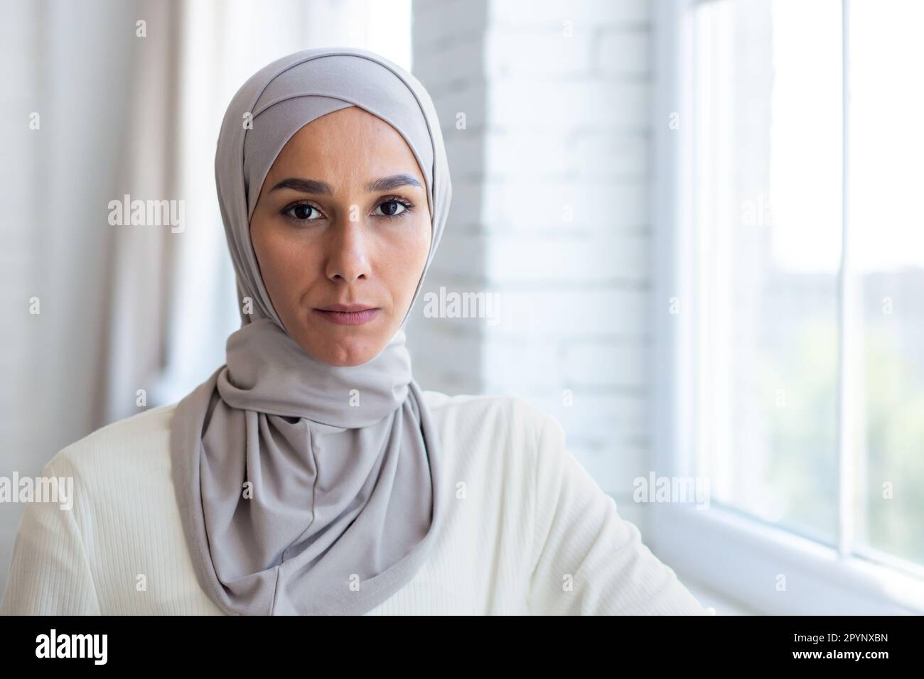 Gros plan. Portrait d'une jeune femme musulmane dans un hijab debout près de la fenêtre à la maison, en toute confiance et sérieusement regardant la caméra. Banque D'Images
