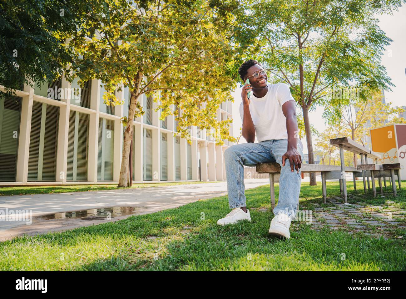 Heureux africain américain étudiant adolescent d'université ayant une conversation d'appel drôle et rire. Portrait d'un jeune homme parlant à l'aide d'un téléphone portable assis sur un banc à l'extérieur sur le campus universitaire. Photo de haute qualité Banque D'Images