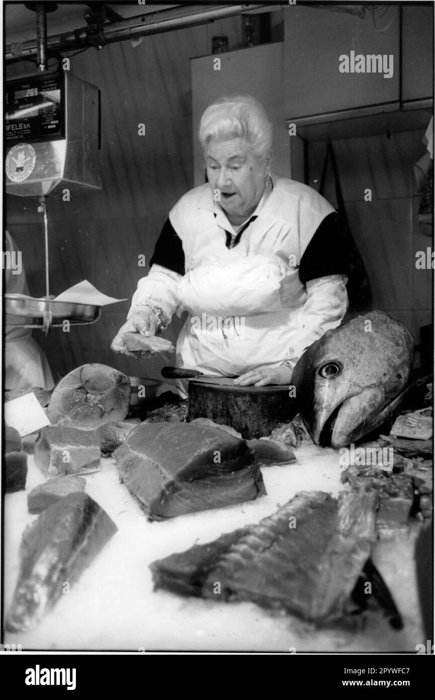 Espagne, Barcelone. Femme de marché avec un tablier blanc se tient derrière son stand de poisson avec de grandes pièces de poisson (thon) et une tête de poisson. Vue de l'intérieur, noir et blanc. Photo, 1993. Banque D'Images