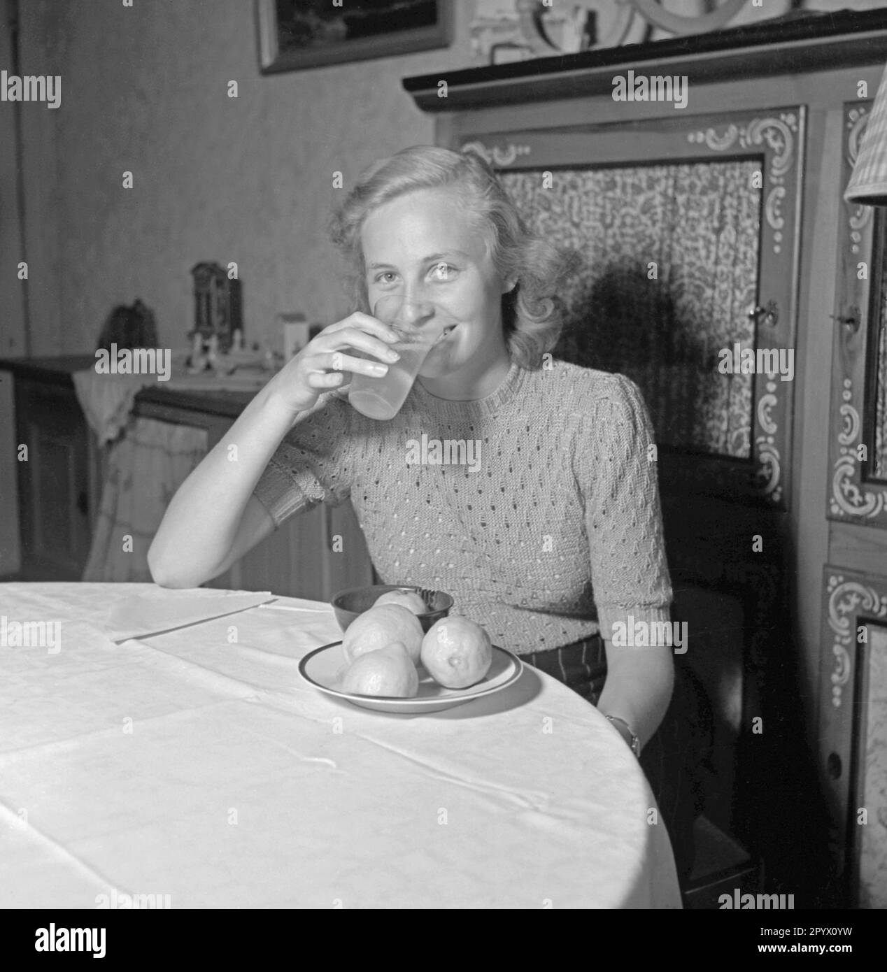 Une jeune femme blonde s'assoit à une table et boit du jus de citron à partir d'un verre, devant elle sur la table se trouve un presse-citron et une assiette avec des citrons. Banque D'Images