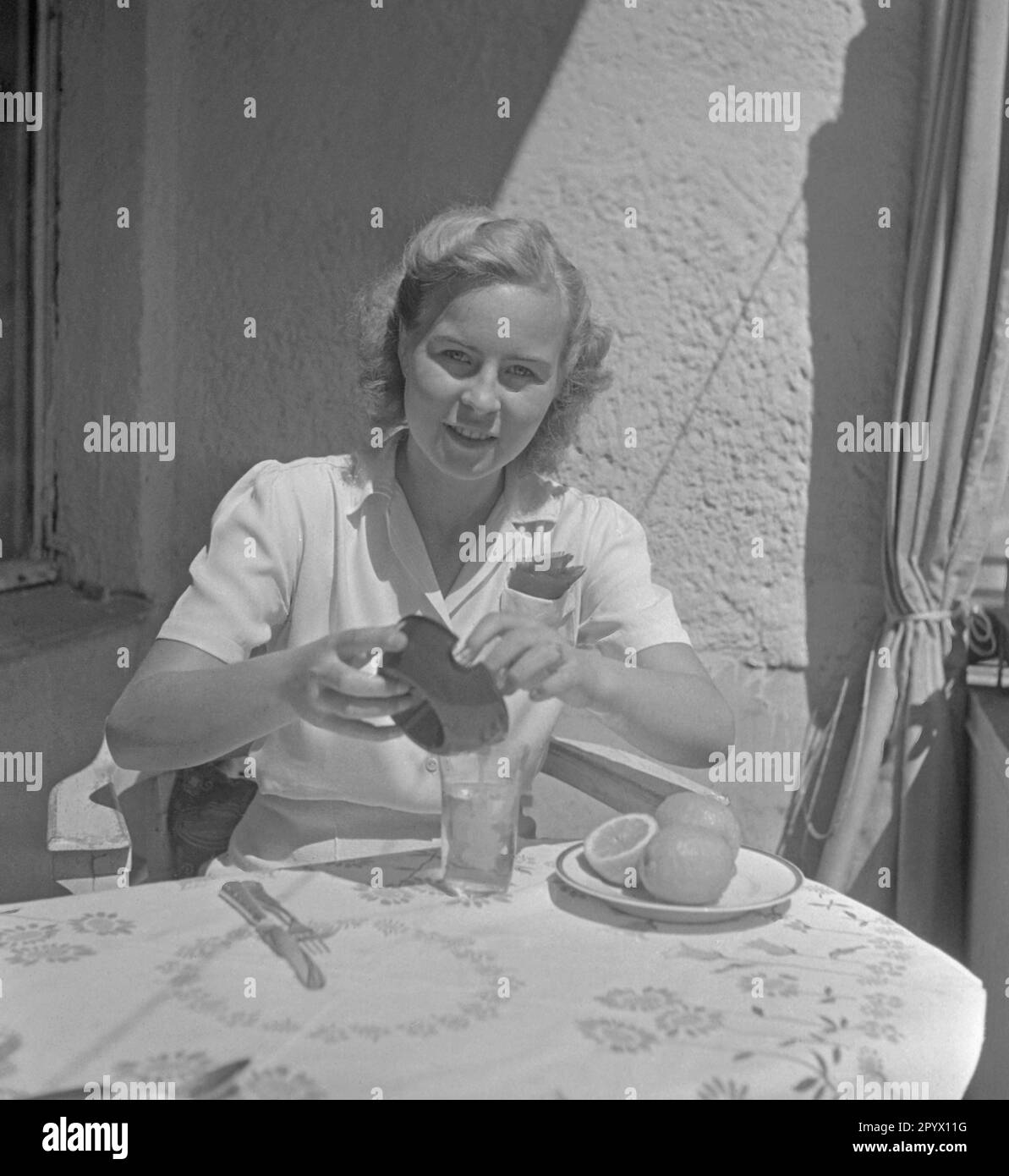 Une jeune femme s'assoit à la table et verse le jus d'un presse-jus de citron dans un verre, devant elle sont des citrons sur une assiette. Banque D'Images