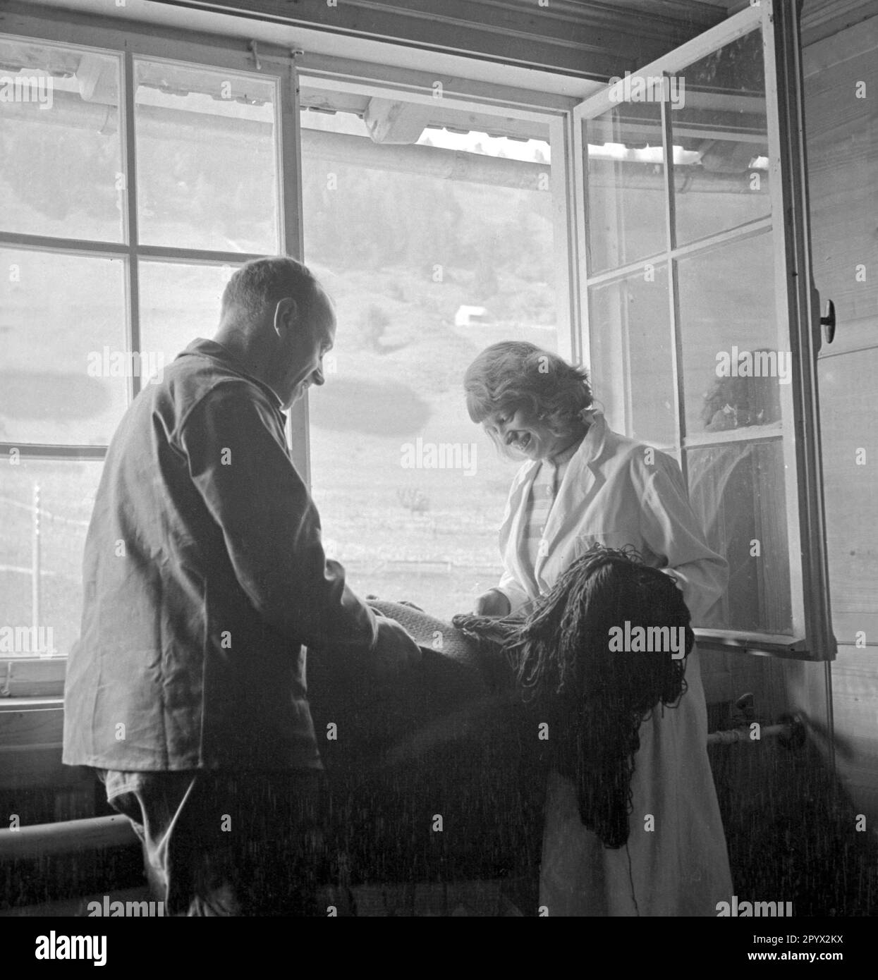 Un homme aide une femme à faire un tapis. Ils sont debout à la fenêtre, en arrière-plan sont les collines d'Allgaeu. Banque D'Images