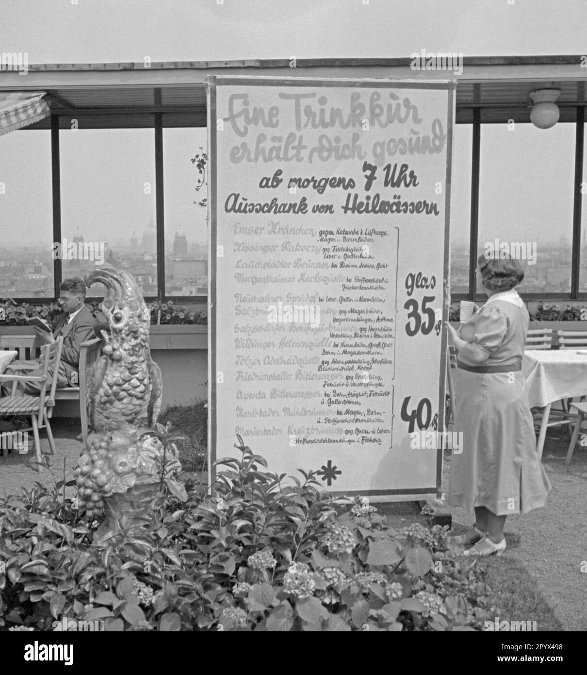 Photo non datée d'une liste de prix pour l'eau curative sur la terrasse du toit du café Europa dans la Maison de l'Europe à Askanischer Platz (Anhalter Bahnhof) à Kreuzberg (Berlin), 1936. Sur le panneau, annonceur d'un remède médical sain d'eau. À droite, un visiteur tient une tasse en porcelaine blanche remplie d'eau curative dans sa main. Sur la gauche, un homme lisant un livre. Au premier plan, un cafatoo assis en pierre. Banque D'Images