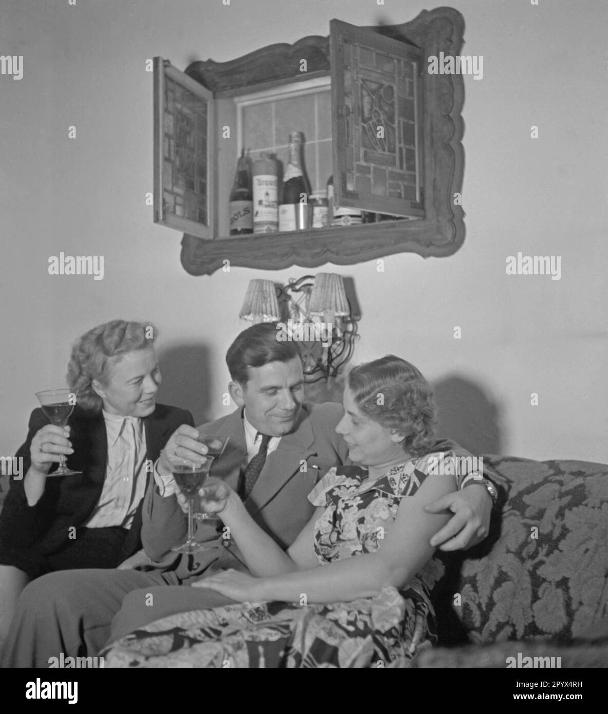 « Un homme boit de l'alcool sur un canapé avec deux femmes. Vous trouverez ci-dessus des bouteilles de la société ''Lucas bols'', une bouteille de champagne et d'autres boissons alcoolisées. Photo non datée, probablement à partir de l'an 1950.' Banque D'Images