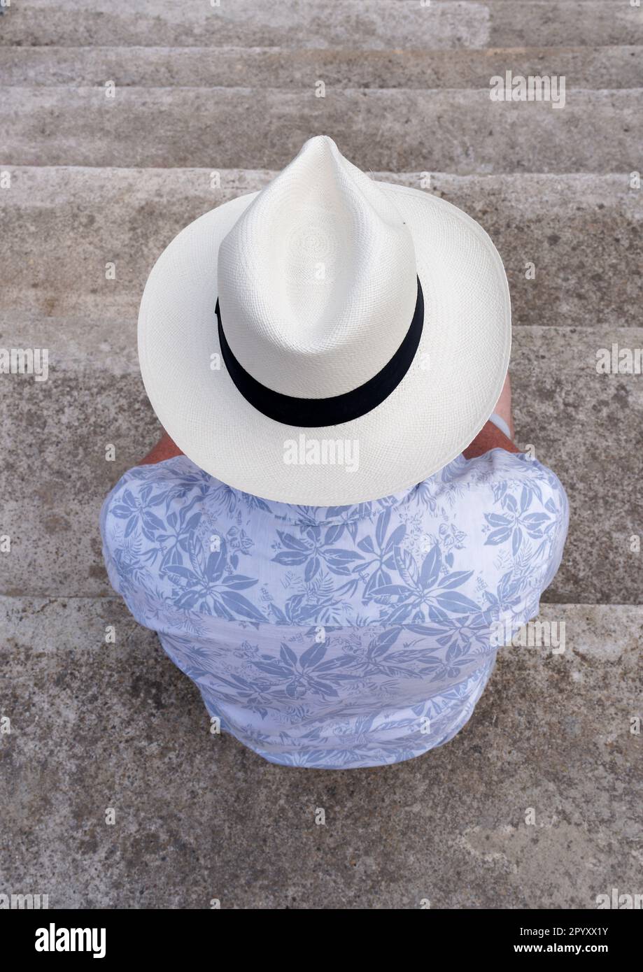 Un homme portant une chemise bleue et un grand chapeau de Panama beige sur sa tête, assis sur quelques marches de pierre et avec son dos à la caméra. Cadix, Espagne Banque D'Images