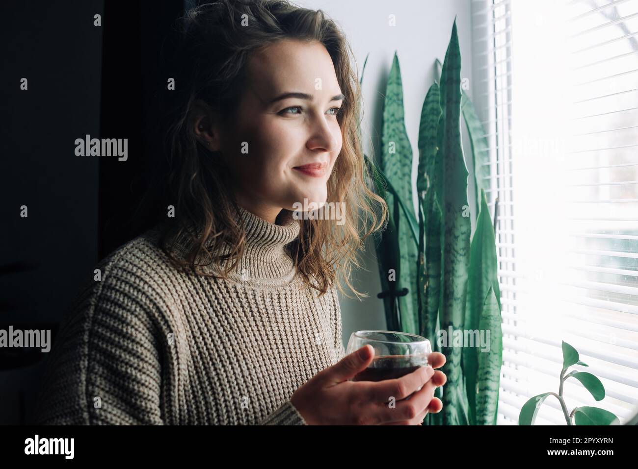 Une jeune femme sereine savoure une tasse de thé chaud tout en admirant le paysage urbain époustouflant d'une fenêtre, en prenant un moment paisible pour se détendre et Banque D'Images