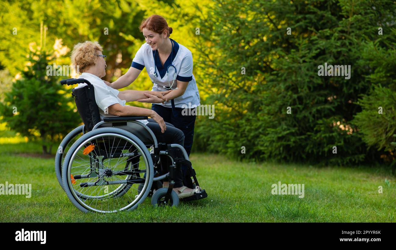 Une infirmière tient une femme caucasienne âgée dans un fauteuil roulant à la main comme support. L'infirmière marche avec un patient dans le parc. Banque D'Images