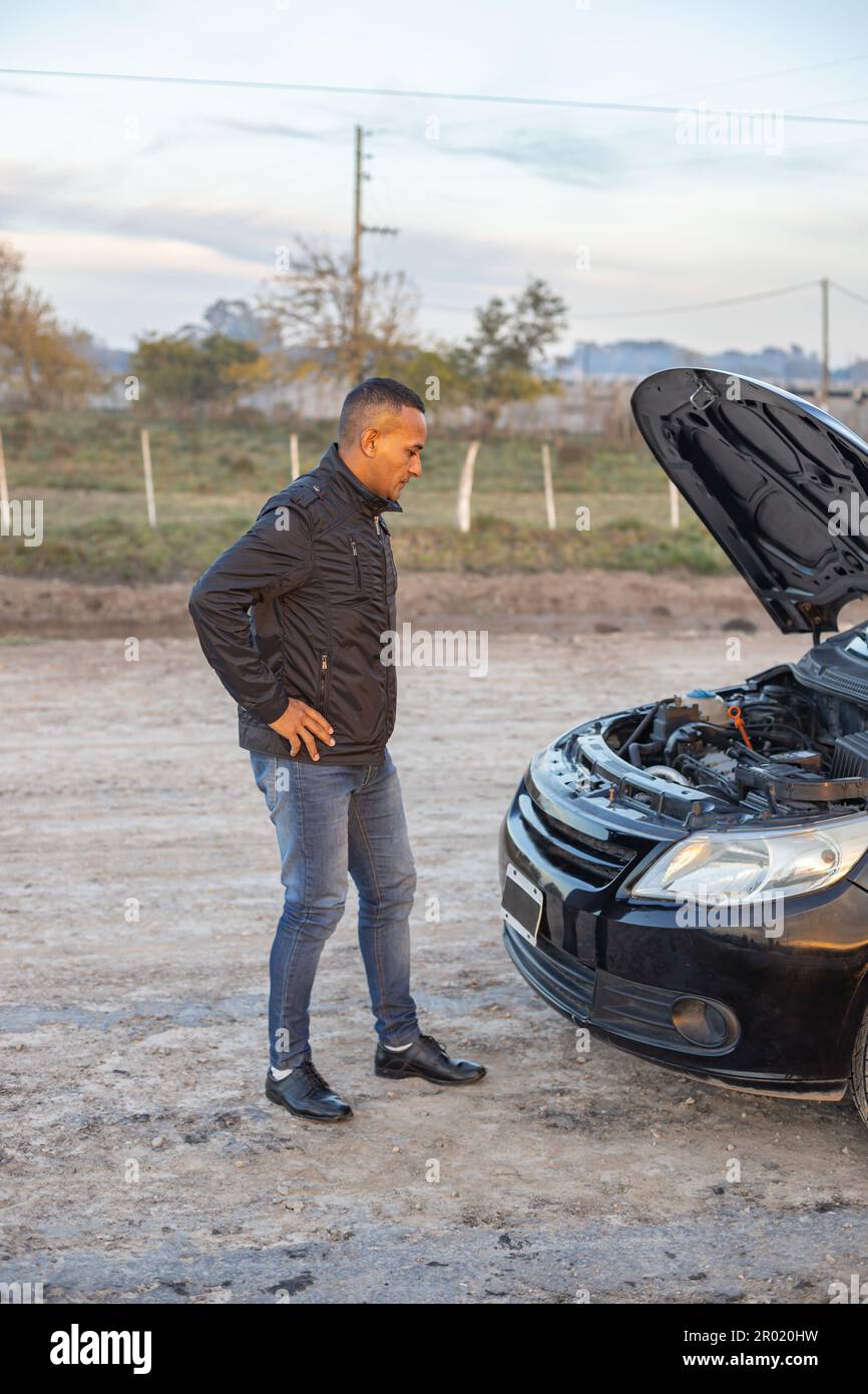 Jeune Latino homme regardant le moteur de sa voiture en panne sur le côté de la route. Banque D'Images