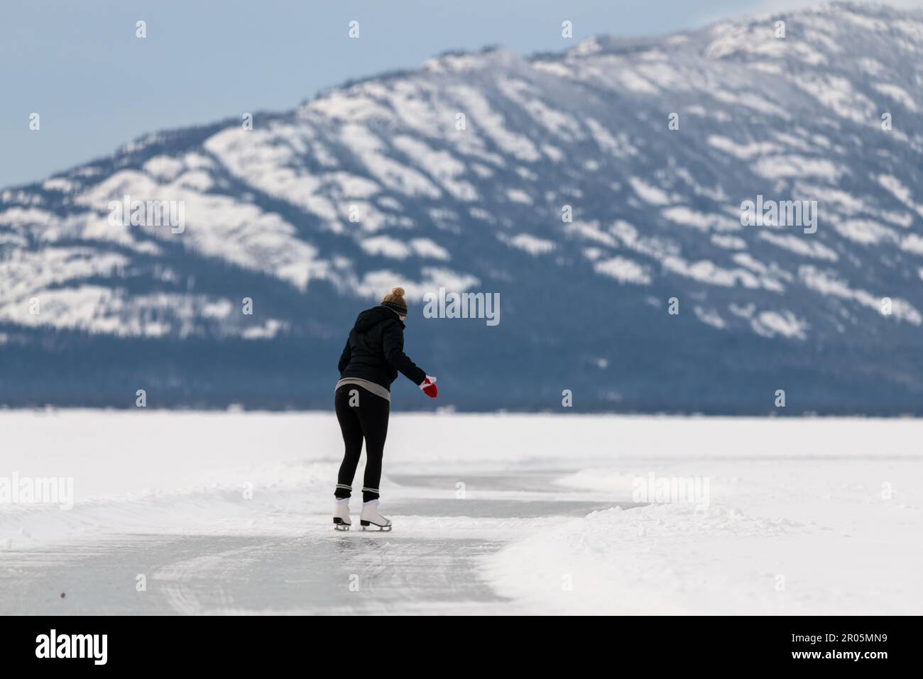 Femme patinant sur un lac gelé dans le nord du Canada pendant l'hiver avec des montagnes enneigées derrière. Vue sur la nature sauvage dans le territoire du Yukon une personne. Banque D'Images