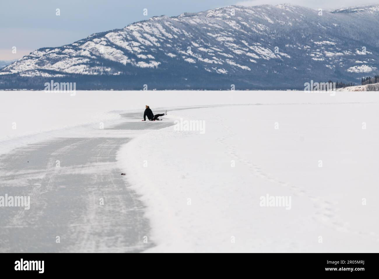 Femme patinant sur un lac gelé dans le nord du Canada pendant l'hiver avec des montagnes enneigées derrière. Vue sur la nature sauvage dans le territoire du Yukon une personne. Banque D'Images