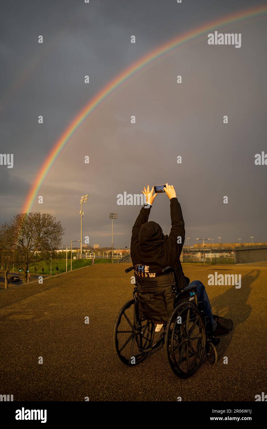 Une femme portant un sweat à capuche noir et assise dans un fauteuil roulant prend une photo d'un arc-en-ciel à l'aide de son téléphone portable. L'emplacement est le Parc olympique de Stratford Banque D'Images