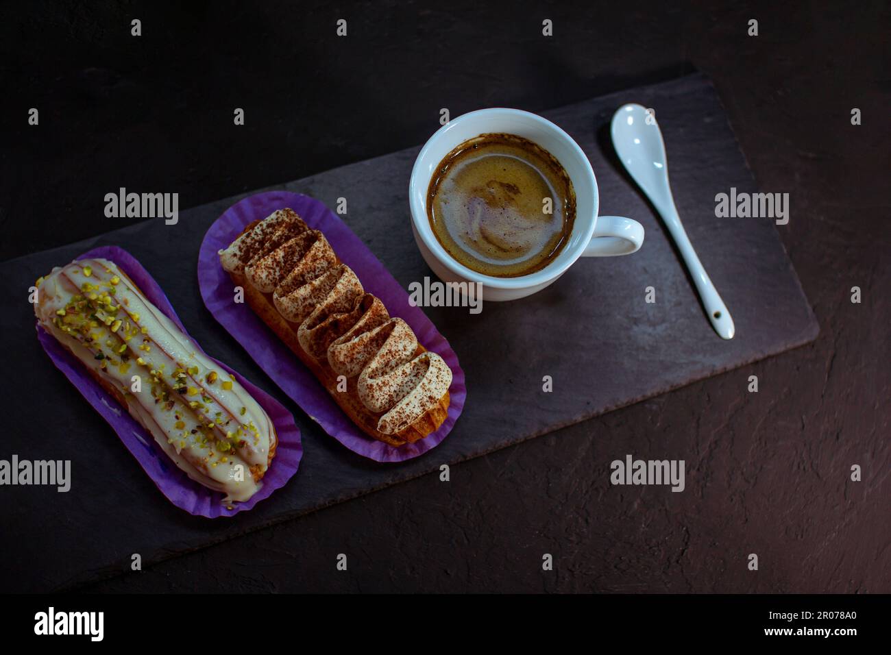 Tasse de café et délicieux gâteaux eclair - tiramisu et pistache aromatisés à la crème, au glaçage et aux noix hachées, sur fond noir. Banque D'Images