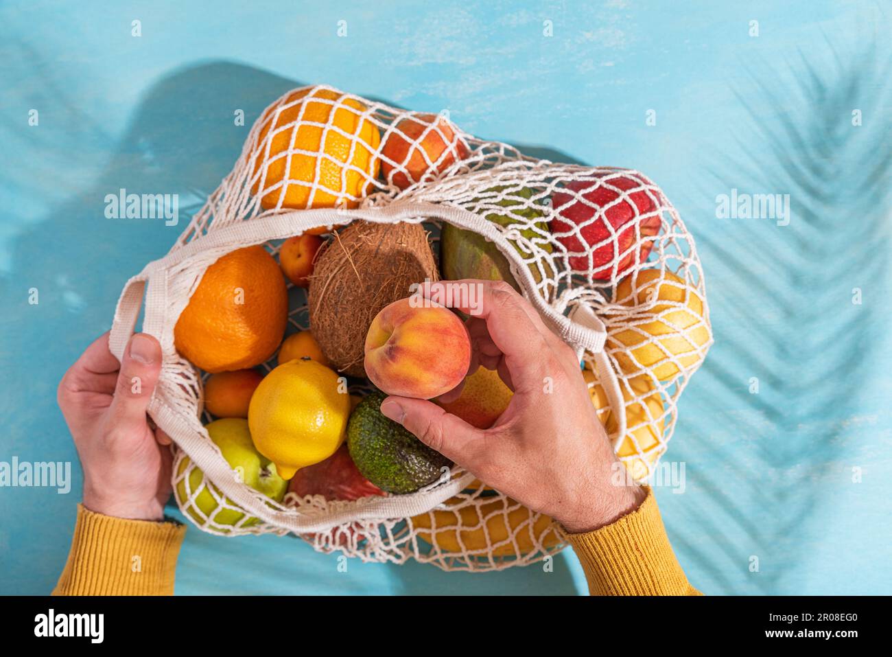 Homme à la main prenant des fruits d'avocat de sac de shopping de filet avec des fruits tropicaux. Arrière-plan bleu avec ombre à la paume Banque D'Images