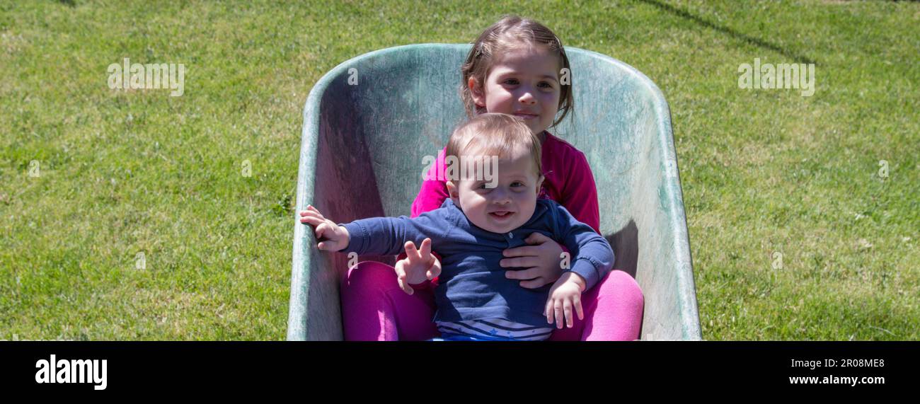 Image de deux adorables enfants souriants se tenant à l'intérieur d'une brouette. Amour fraternel comme ils jouent dans le jardin avec leur soeur de fabrication Banque D'Images