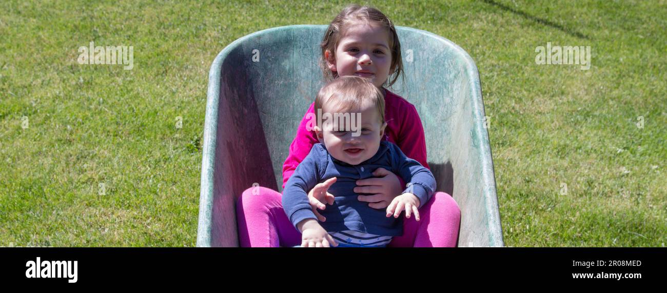 Image de deux adorables enfants souriants se tenant à l'intérieur d'une brouette. Amour fraternel comme ils jouent dans le jardin avec leur soeur de fabrication Banque D'Images