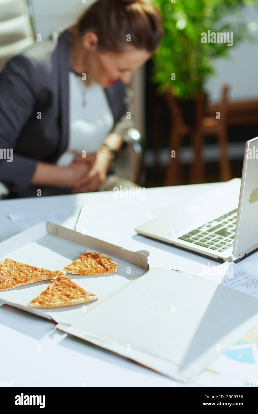 Un lieu de travail durable. Gros plan sur une femme employée dans un bureau vert avec une pizza et un ordinateur portable ayant des douleurs à l'estomac. Banque D'Images