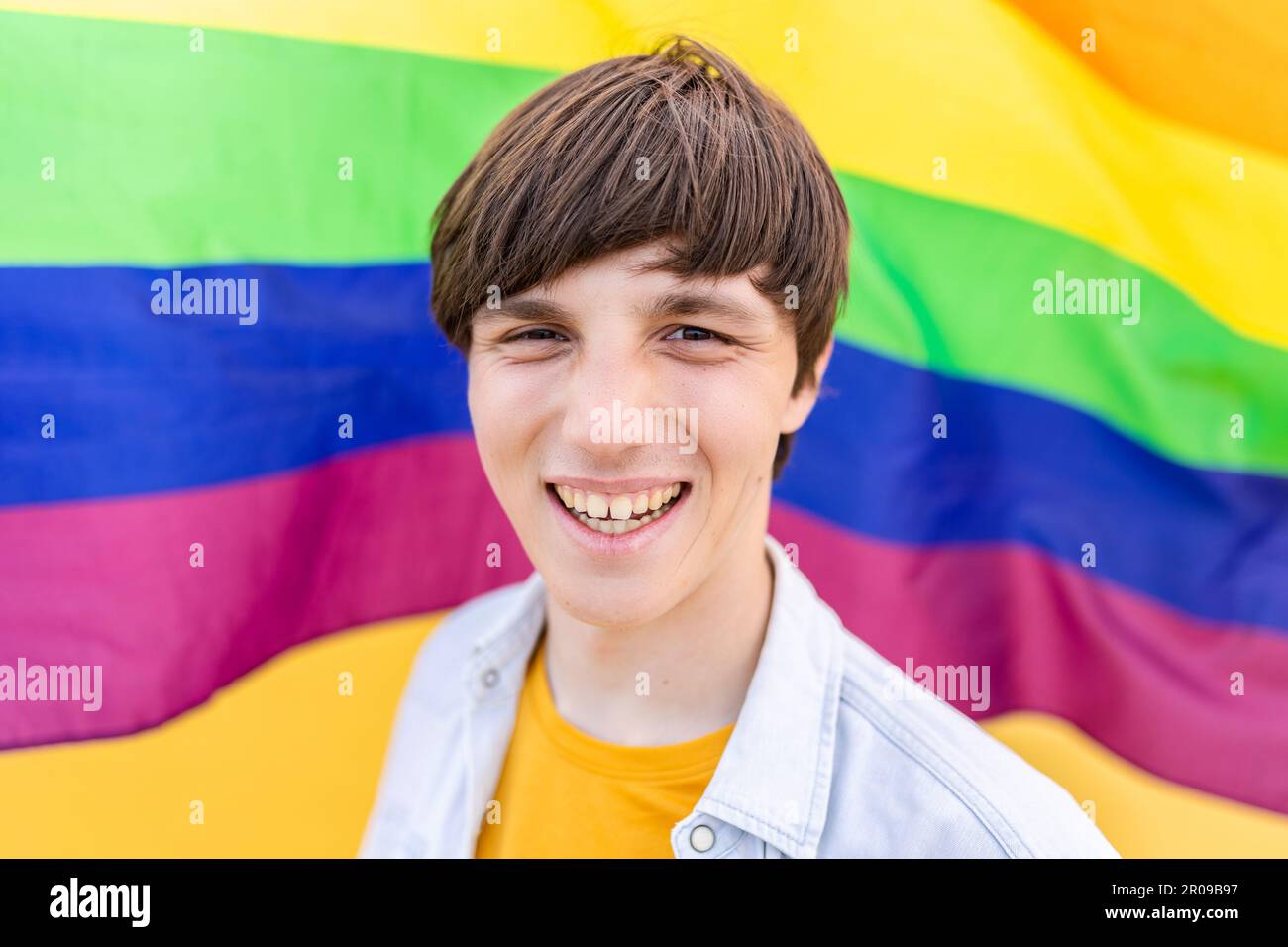 Jeune homme gay souriant à la caméra sur fond de drapeau arc-en-ciel. Banque D'Images