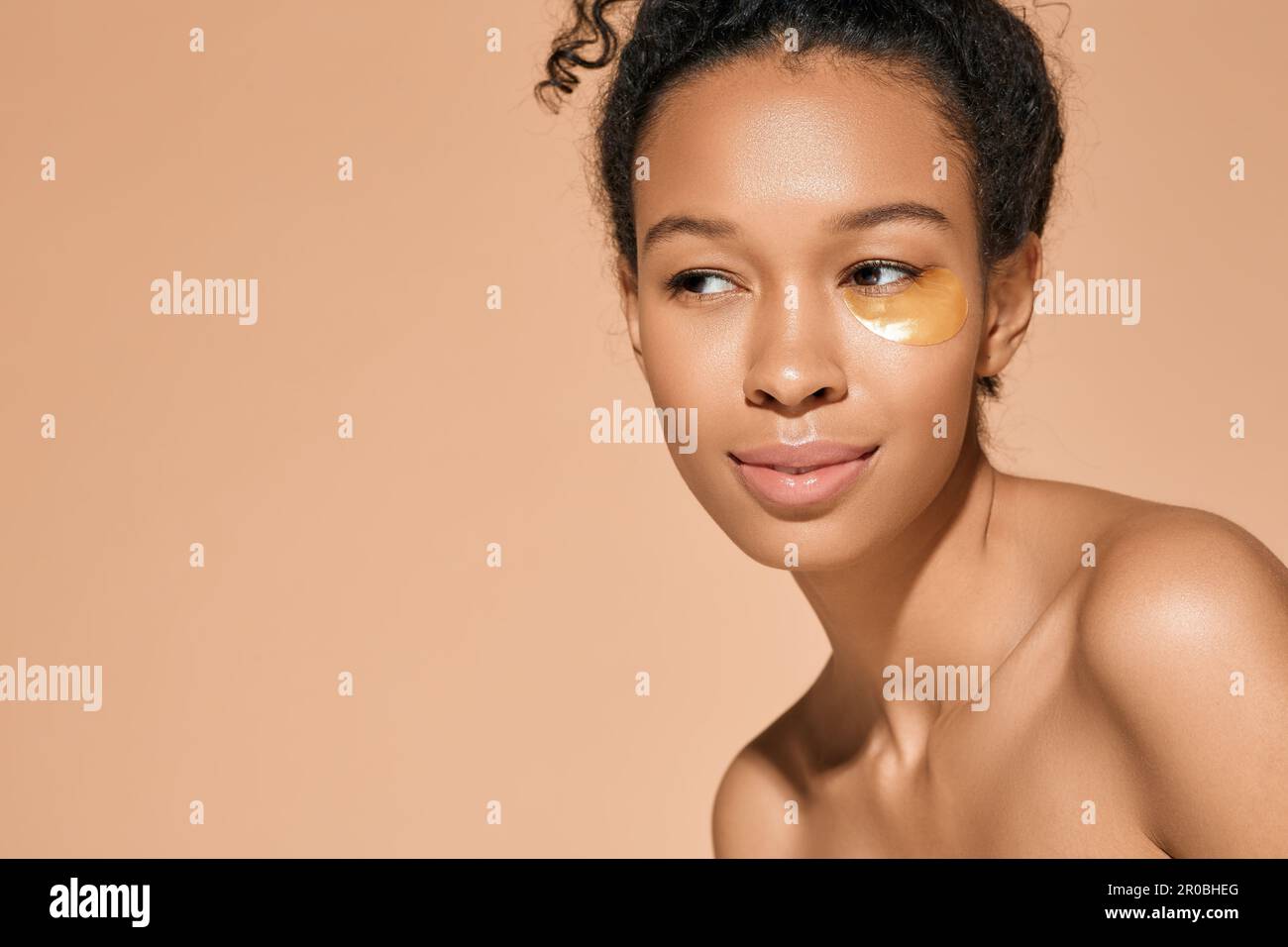Femme afro-américaine avec des taches d'hydrogel dorées sous les yeux pour soulager la puffiness pendant les procédures cosmétiques, isolée sur fond beige. Peau Banque D'Images