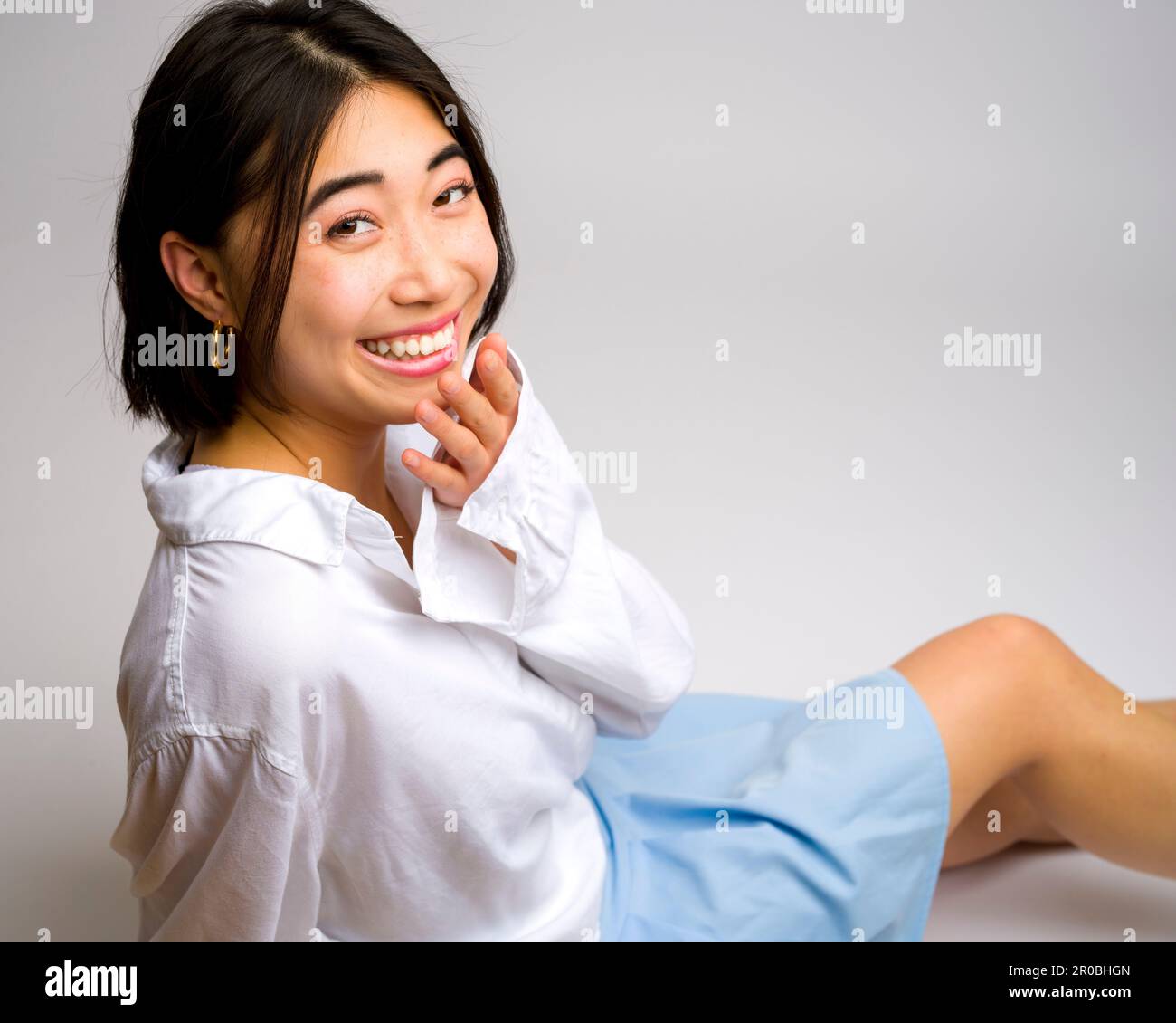 Smiling Young Asian Woman assis sur fond blanc jupe courte manches longues | vue latérale | regarder l'appareil photo Banque D'Images